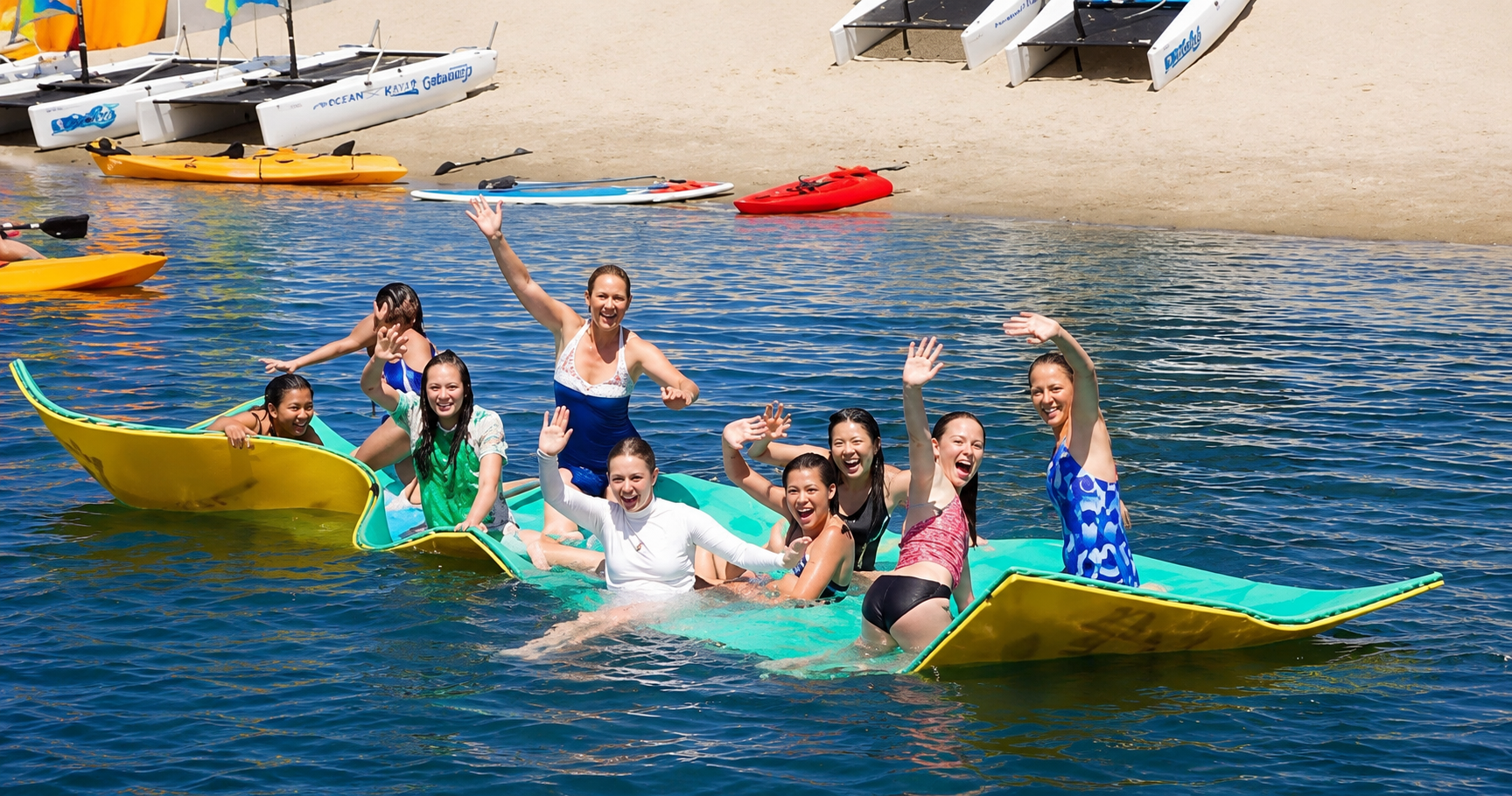 Group relaxing and waving together on Aqua Lily Pad floating mats at Mission Bay Sportcenter